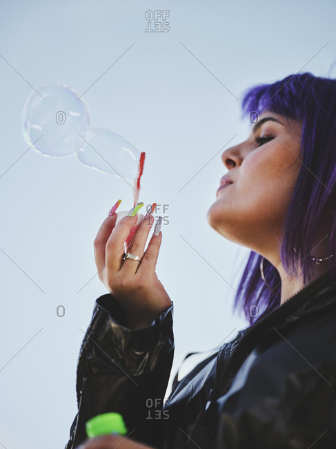From below side view of fashion woman with purple hair smiling and blowing bubbles holding bottle while looking away with manicured hands in bright day
