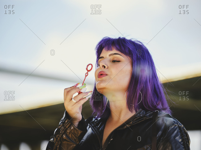 Fashion woman with purple hair smiling and blowing bubbles holding bottle with closed eyes with manicured hands in bright day