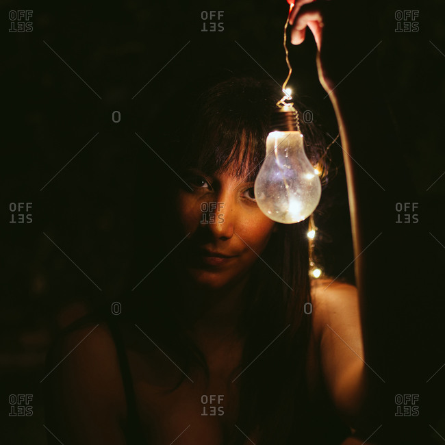 Focused young female model with long dark hair looking at camera and holding burning bulb in darkness