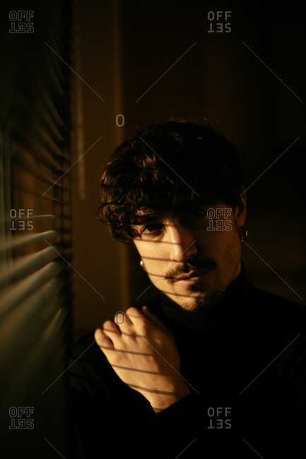 Young melancholic guy in black turtleneck standing next to window with shutters with shadow on face looking at camera