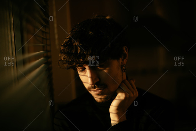 Young melancholic guy in black turtleneck standing next to window with shutters with shadow on face