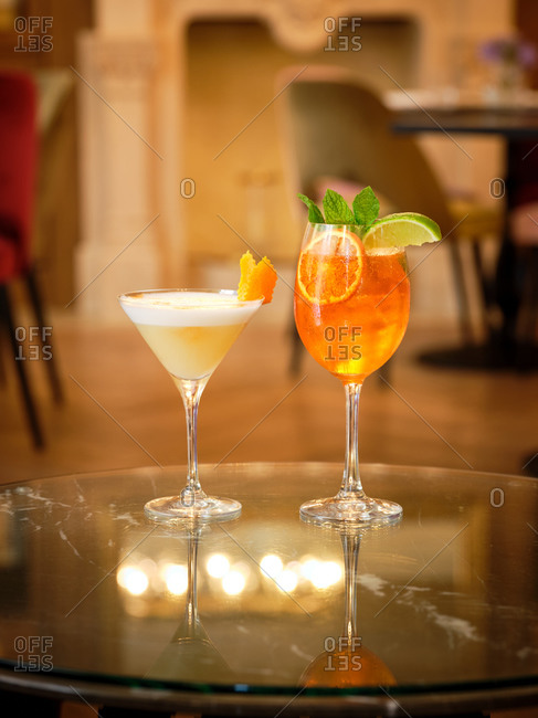 Cocktail of yellow liquid and white foam decorated with orange peel and orange cocktail with ice decorated with mint and lime on glasses table