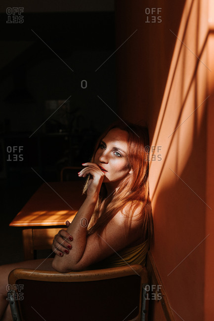 Side view of sensual thoughtful redhead female model in stylish yellow dress with makeup sitting with crossed legs on chair and looking away under beam of sun in dark room
