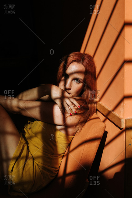 Sensual thoughtful redhead female model in stylish yellow dress with makeup sitting on chair and looking at camera under beam of sun in dark room