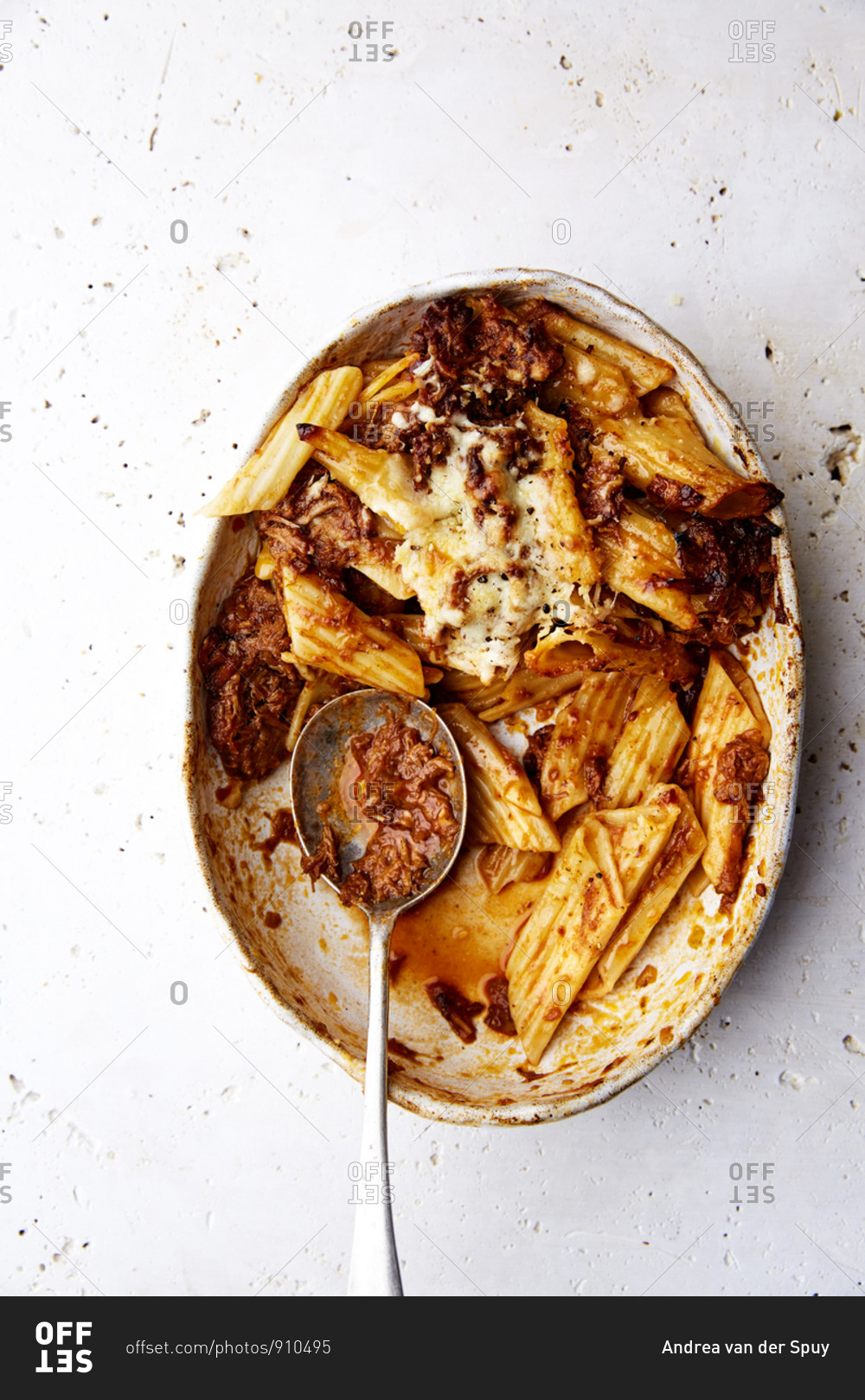 A serving bowl of baked ragu and penne pasta with a silver spoon on a white countertop,