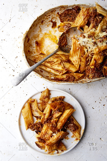 A serving bowl of baked ragu and penne pasta with a plate served on the side on a white countertop,