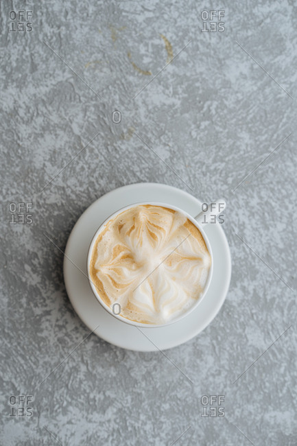Top view of cup of hot cappuccino with latte art served on white saucer on light grey textured background
