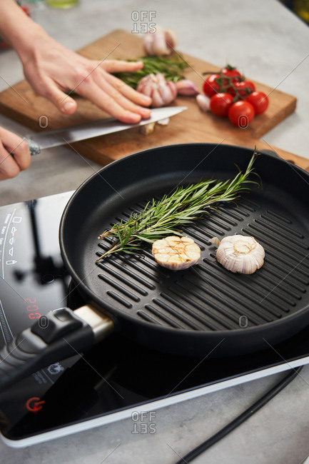 Grill pan with sprigs of fresh rosemary and cut garlic head placed on stove and crop hands of woman crushing garlic on wooden cut board in background