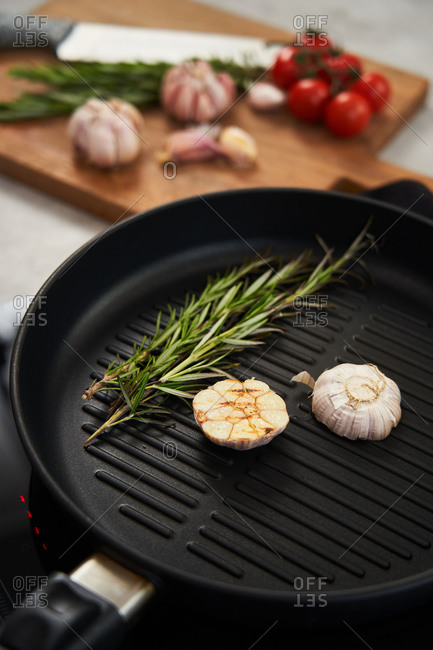 Aromatic grilled sprigs of rosemary and cut in half garlic head on grill pan placed on stove with cutting board with fresh herbs and vegetables in background