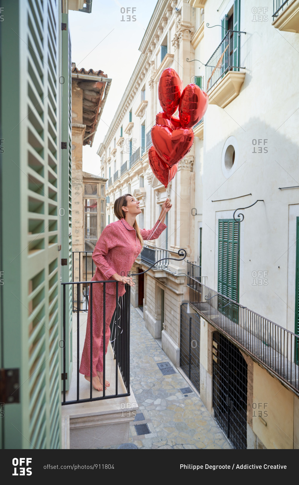Happy lady in pink sleepwear with vivid red heart shape balloons standing on balcony leaning on metal fence and looking up against facades of buildings in old town