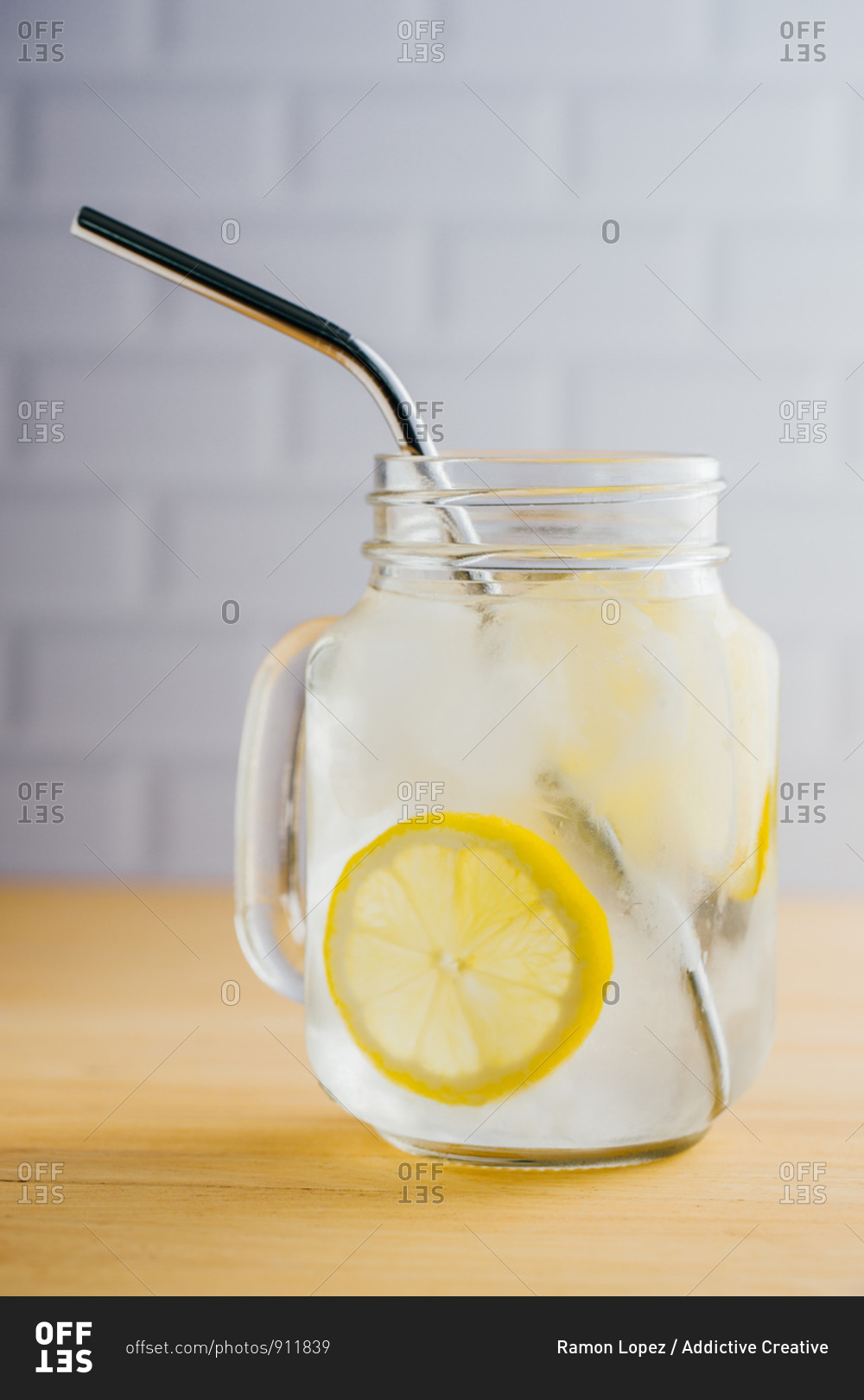 Metallic reusable straw and glass jug with ice and lemon slices on wooden table in kitchen