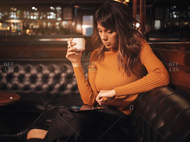 Stylish smart thoughtful woman surfing tablet comfortably sitting on black leather couch in cafe using tablet and drinking coffee in a mug