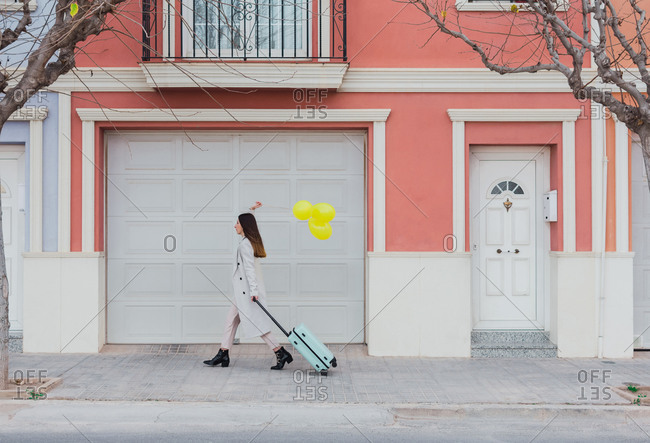 Side view of young stylish female with yellow balloons and suitcase walking on city street next to old styled colorful building