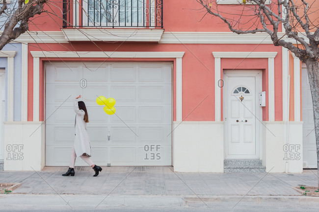 Side view of unrecognizable stylish female covering face with yellow balloons walking with suitcase on city street next to old styled colorful building