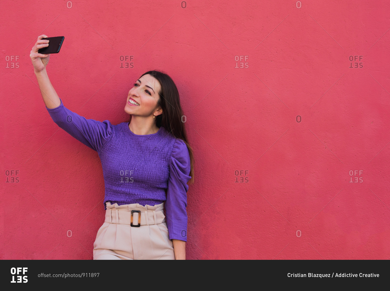 Positive young female in violet blouse and light beige pants taking a selfie on mobile phone while standing against colorful red wall background