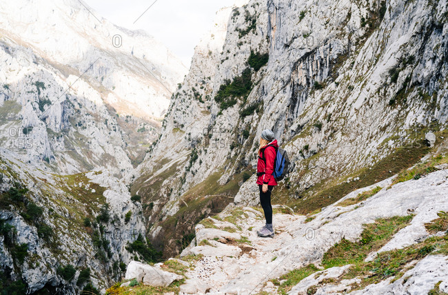 Side view of unrecognizable active woman hiker in red jacket with heavy backpack looking up at mountain in peaks of Europe, Asturias, Spain