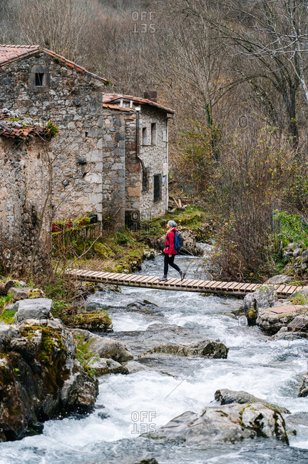 From above active woman tourist in red jacket with heavy backpack crossing way on wooden bridge in village in peaks of Europe, Asturias, Spain