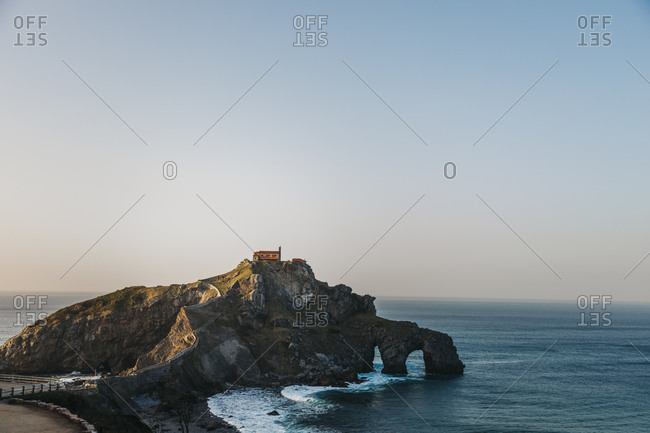 Paving stone way leading along stone bridge and ridge of rocky hill to lonely country house on island Gaztelugatxe surrounded by tranquil sea water with white foam waves