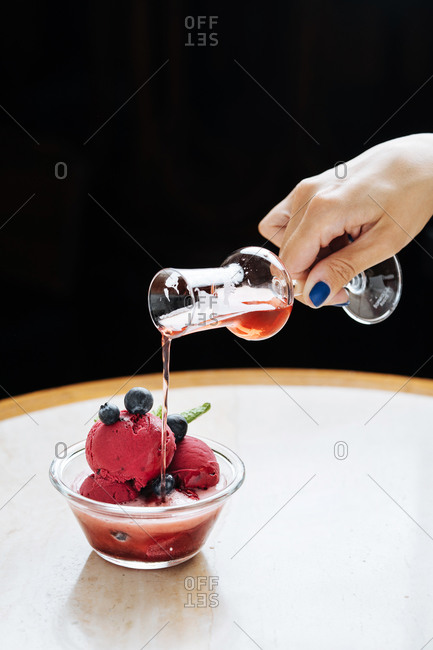 Cropped unrecognizable woman hand pouring a red beverage from a glass on purple scoops of ice cream in glass bowl decorated with fresh blueberry and mint and served in restaurant