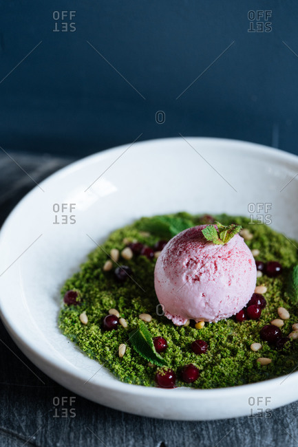 From above of scoop of purple ice cream on green mousse decorated with nuts and fresh mint in white bowl