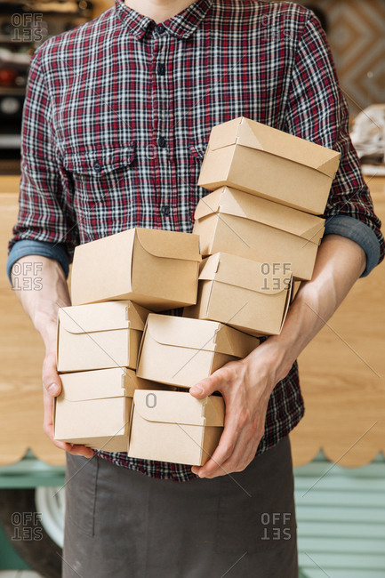 Unrecognizable man in plaid shirt holding lots of small cardboard boxes with delivered food in restaurant