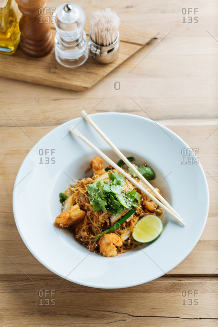 From above of tasty noodles with fried shrimps slices of lime and fresh parsley in plate with wooden chopsticks