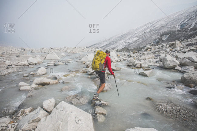 Backpacker uses large boulders to cross river next to glacier.