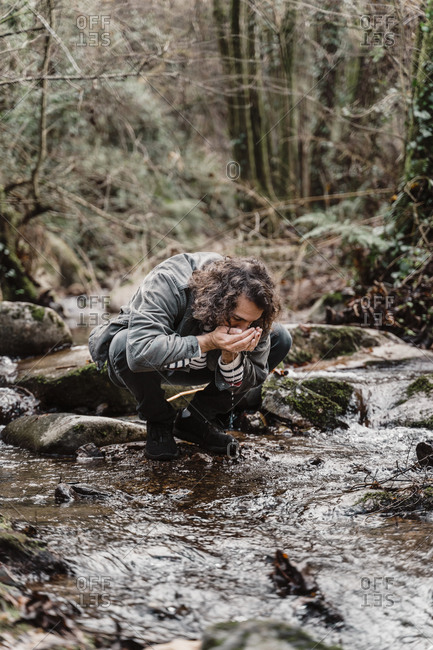 Young man crouching at stream in forest- drinking water from cupped hands