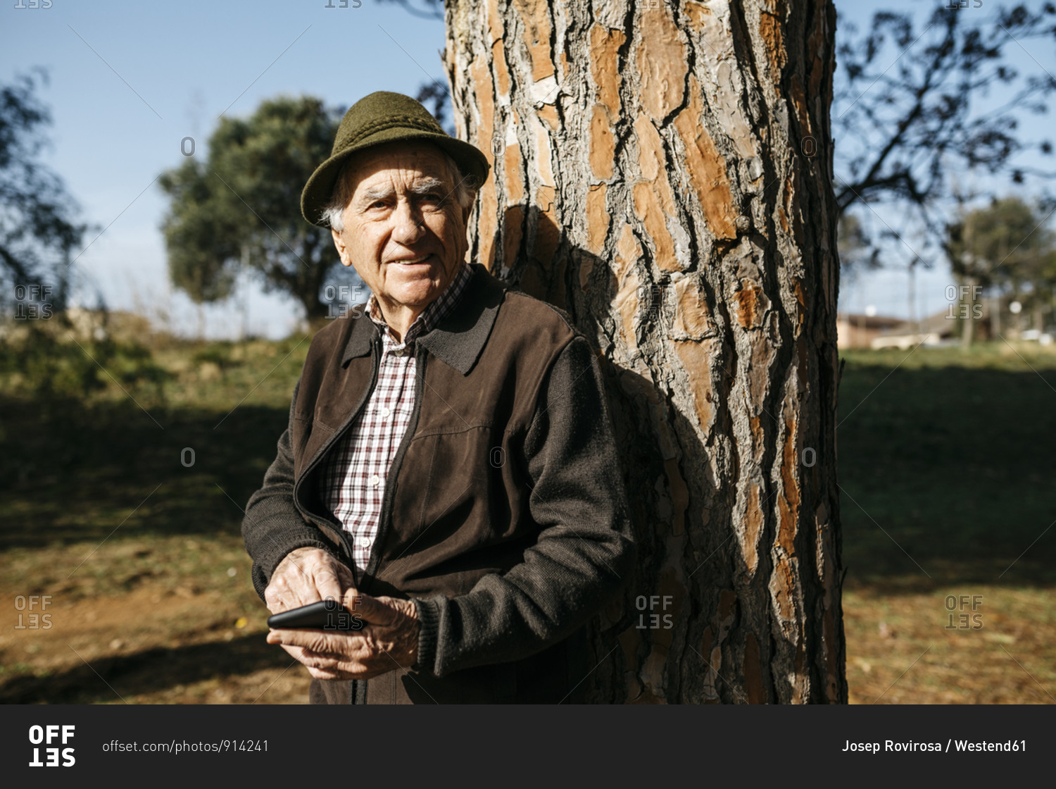 Old man using smartphone- leaning on tree trunk