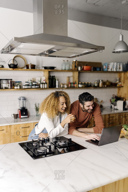 Stock photo of a middle aged woman and man standing in a kitchen and looking at a laptop. They are smiling.