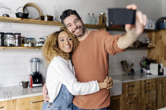 Stock photo of a middle aged woman and man standing in a kitchen and laughing. They are taking a selfie together.