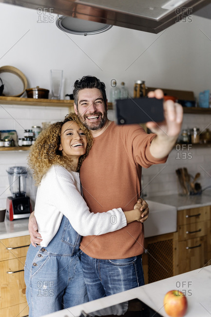 Stock photo of a middle aged woman and man standing in a kitchen and laughing. They are taking a selfie together.