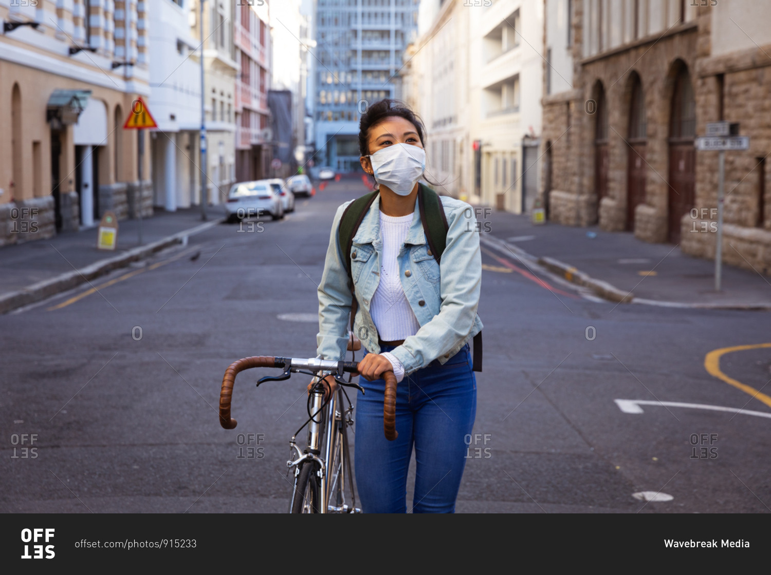 Front view of a mixed race woman with dark hair out and about in the city streets during the day, wearing a face mask against air pollution and coronavirus, walking with her bicycle with buildings in the background.