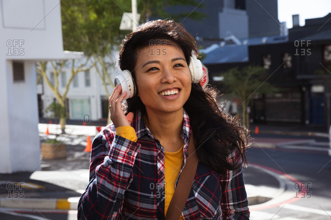 Front view of a mixed race woman with long dark hair out and about in the city streets during the day, wearing headphones, a checked shirt and walking in a city street with buildings in the background.