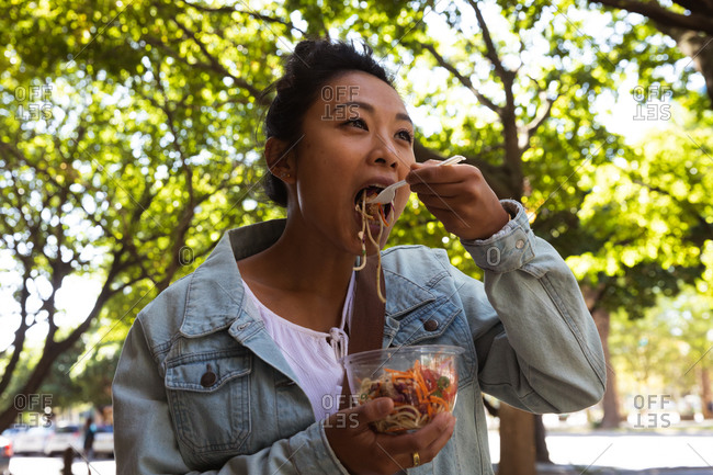 Low angle front view of a mixed race woman with long dark hair out and about in the city streets during the day, eating takeaway lunch, wearing a denim jacket and walking in a city street with trees in the background.