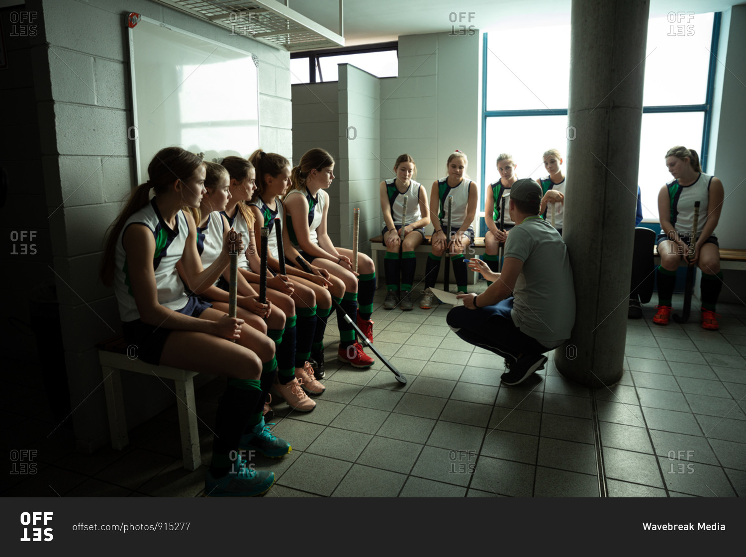 Side view of a Caucasian male field hockey coach interacting with a group of female Caucasian field hockey players, sitting in a changing room, showing them a game plan