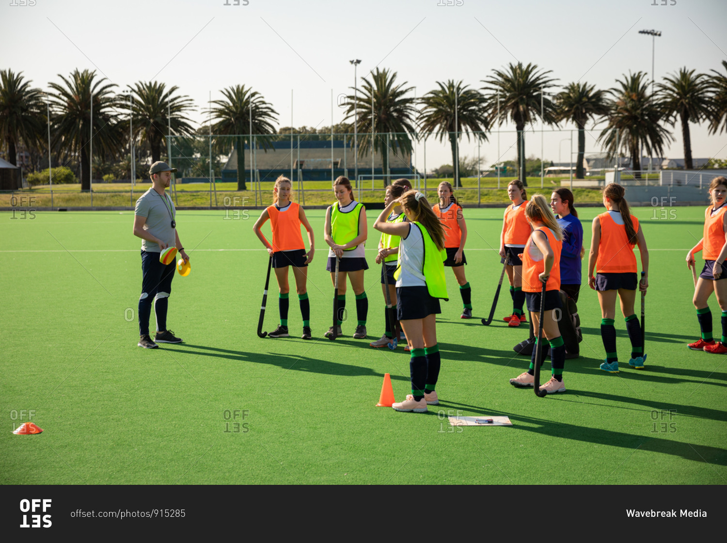 Side view of a group of female Caucasian field hockey players and their Caucasian male field hockey coach, training before a game, working out on a field hockey pitch, doing exercises, preparing for shooting training on a sunny day