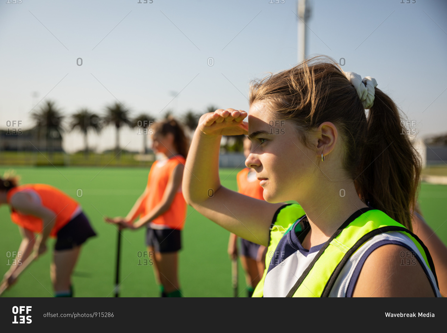 Side view of a Caucasian female field hockey player training before a game, working out on a hockey pitch, shading her eyes from the sun, with her teammates in the background, on a sunny day