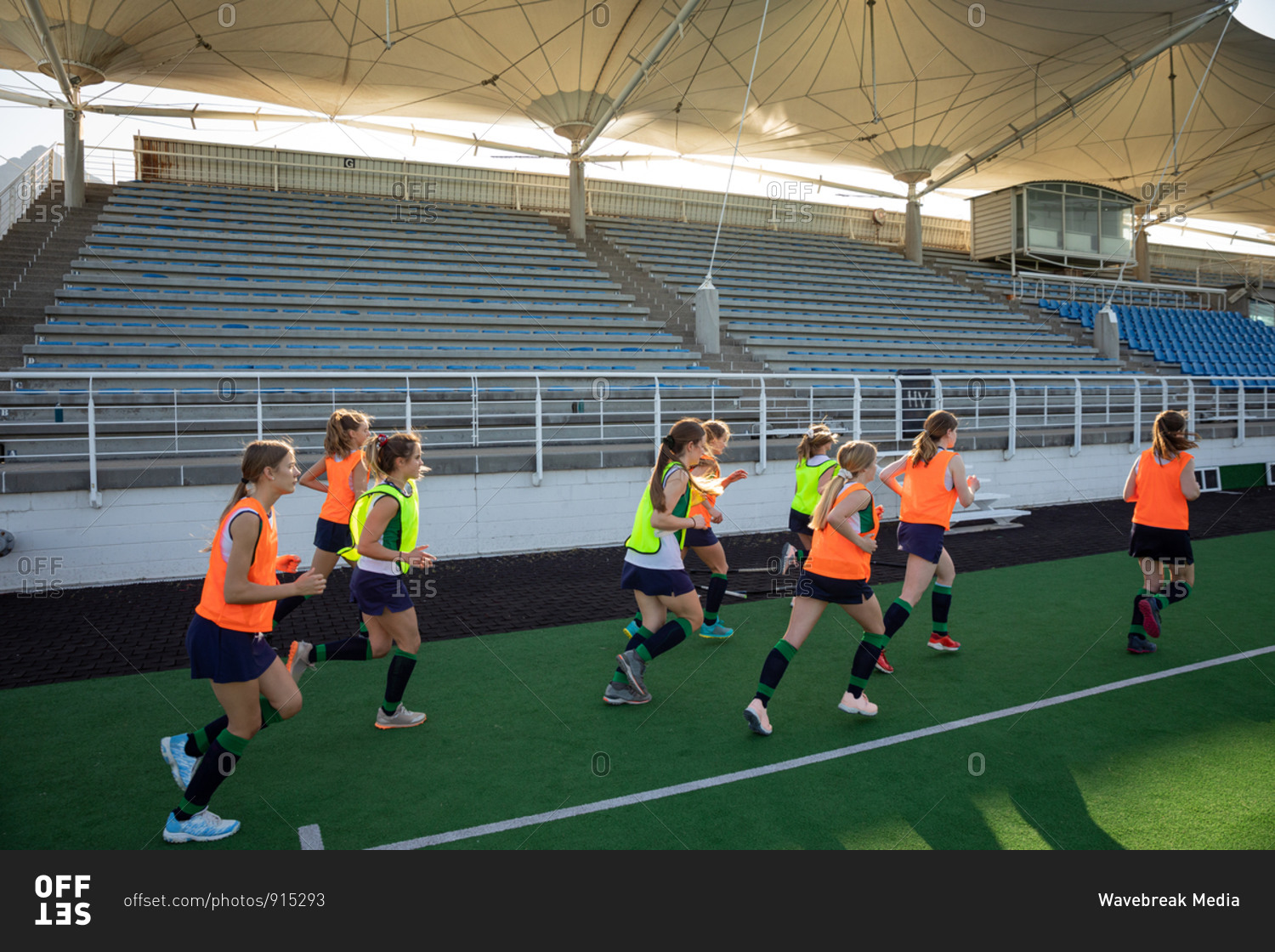 Side view of a group of female Caucasian field hockey players and their Caucasian male field hockey coach, training before a game, working out on a field hockey pitch, doing exercises, running, on a sunny day