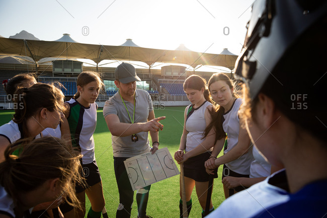 Group of female Caucasian field hockey players huddling around their Caucasian male coach who gives them game plan and points before a game on a sunny day