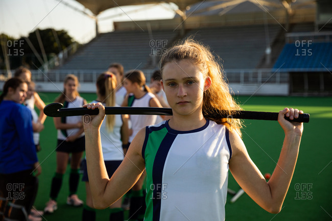Portrait of a Caucasian female field hockey player standing on a hockey pitch with a hockey stick on her shoulder looking at camera with her teammates standing in a row behind her