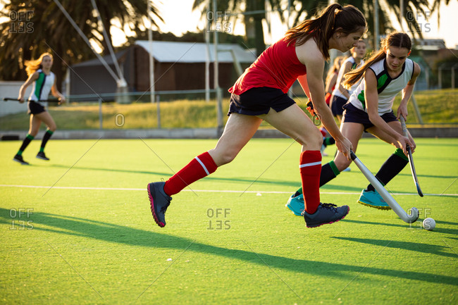 Side view of a Caucasian female field hockey player, during a field hockey game, running with a ball, holding a hockey stick, with her teammates and opponents in the background, on a sunny day
