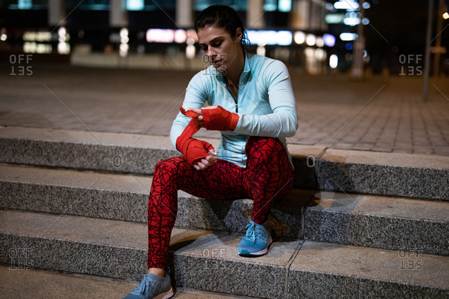 Front view of a fit Caucasian woman with long dark hair wearing sportswear exercising outdoors in the city in the evening, sitting on steps, wrapping her hands in boxing tape with buildings in the background