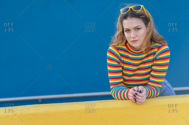 Young woman dressed in a colored striped jersey and yellow glasses on a blue background leaning against a yellow wall