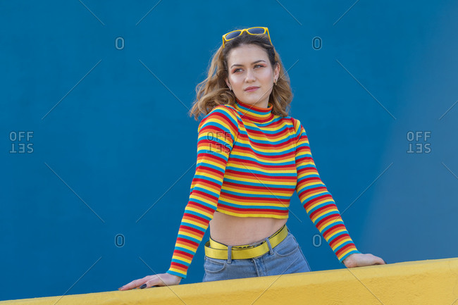 Young woman dressed in a colored striped jersey and yellow glasses on a blue background leaning against a yellow wall
