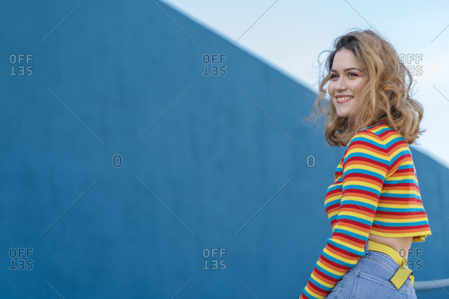 Happy young woman dressed in a colored striped jersey and yellow glasses on a blue background sitting on her back on a yellow wall