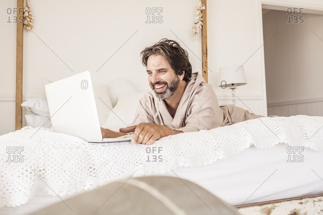 Man wearing bathrobe and lying on bed in bedroom- using laptop