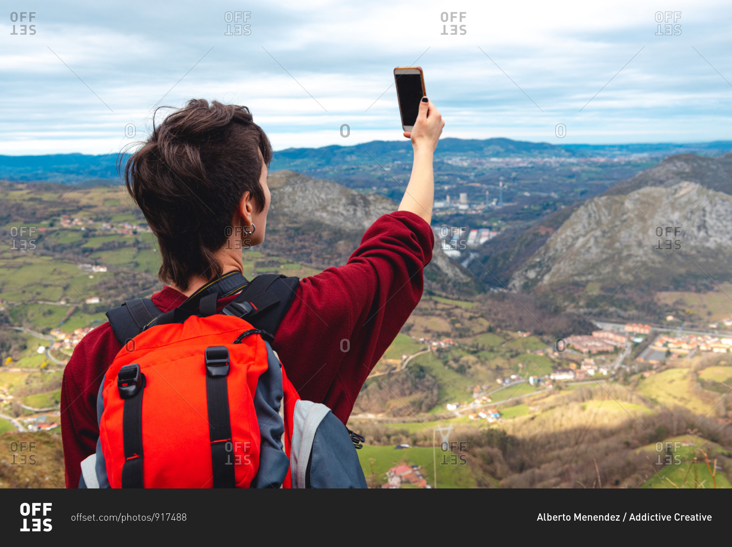 Back view of unrecognizable woman with backpack standing with arm raised and taking shot with smartphone of wonderful scenery with small villages and town in valley against foggy ridges at horizon under cloudy sky in Asturias