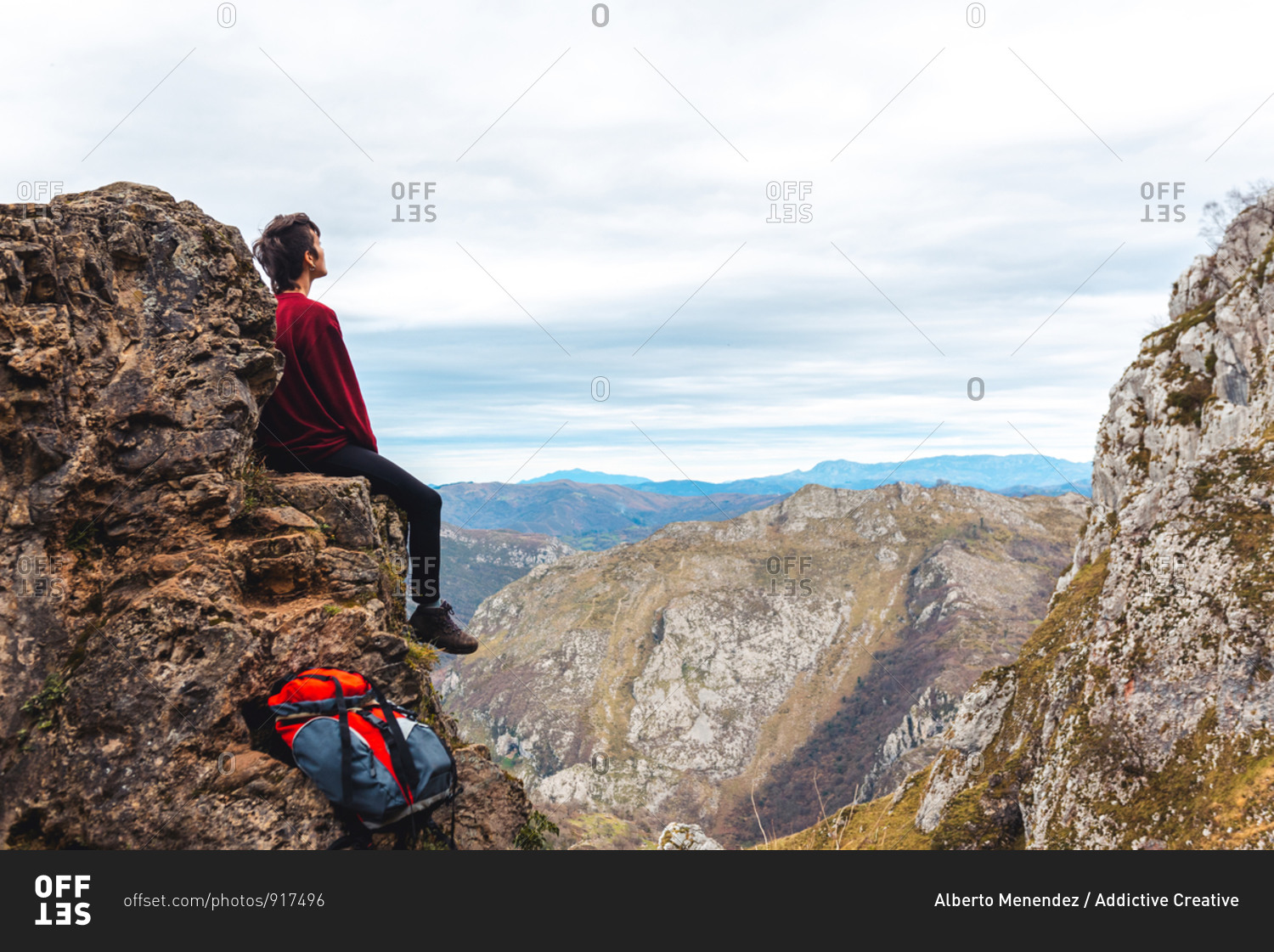 Side view of tourist sitting on edge of cliff enjoying freedom and admiring amazing scenery of countryside located in valley at mountain foothill against foggy forested hills and plain under sky with lush gray clouds in Spain