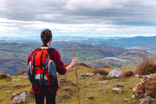 Back view of unrecognizable hiker with backpack and stick standing with arms outstretched and enjoying freedom viewing majestic scenery of countryside located along river shore in valley against foggy ridges at horizon under cloudy sky in Spain